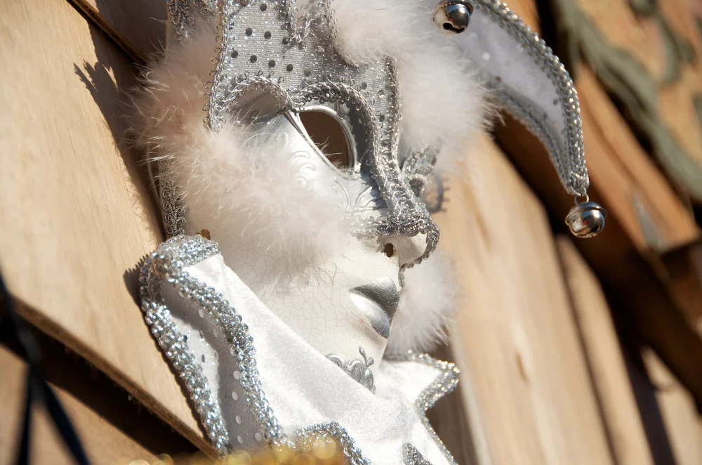 Ornate silver and white jester-style masquerade mask with feathers and small bells resting on a warm wooden surface