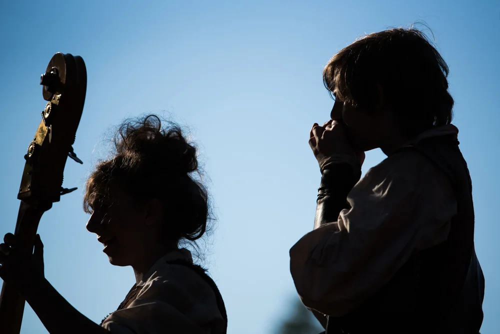 A wide silhouette photograph of two musicians against a soft dusk sky, one holding a stringed instrument and the other a wind instrument, rendered in deep indigo and pale blue with a quiet, communal mood.